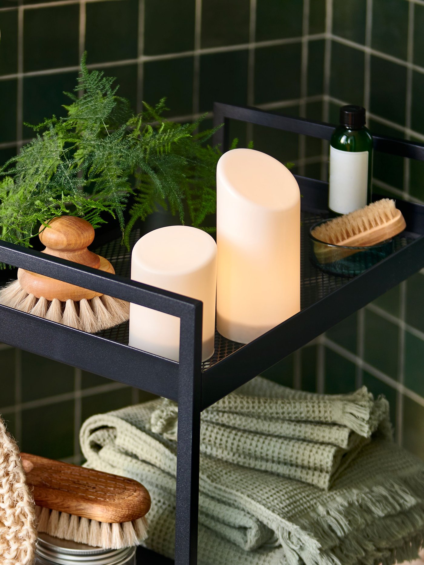 Ein schwarzer NISSAFORS-Trolley, gefüllt mit Spa-Essentials wie grünen Handtüchern, Körperbürsten und LED-Blockkerzen von ÄDELLÖVTRÄD.
