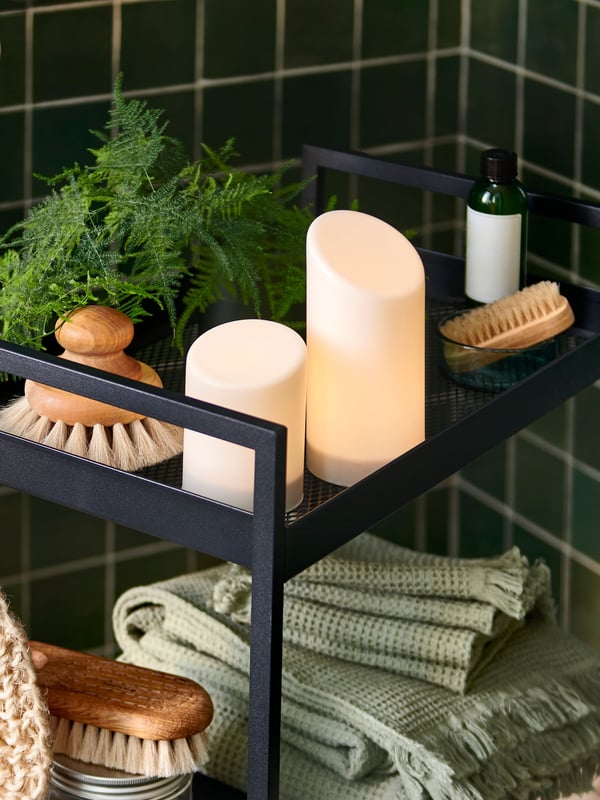 Ein schwarzer NISSAFORS Rollwagen, gefüllt mit Spa-Essentials wie grünen Handtüchern, Körperbürsten und ÄDELLÖVTRÄD LED-Stumpenkerzen.