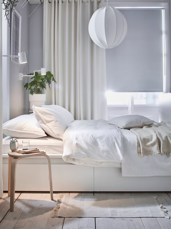 A white MALM bed and two MALM chests with 6 drawers behind it. Two MULIG clothes bars are on the wall holding bright clothes.