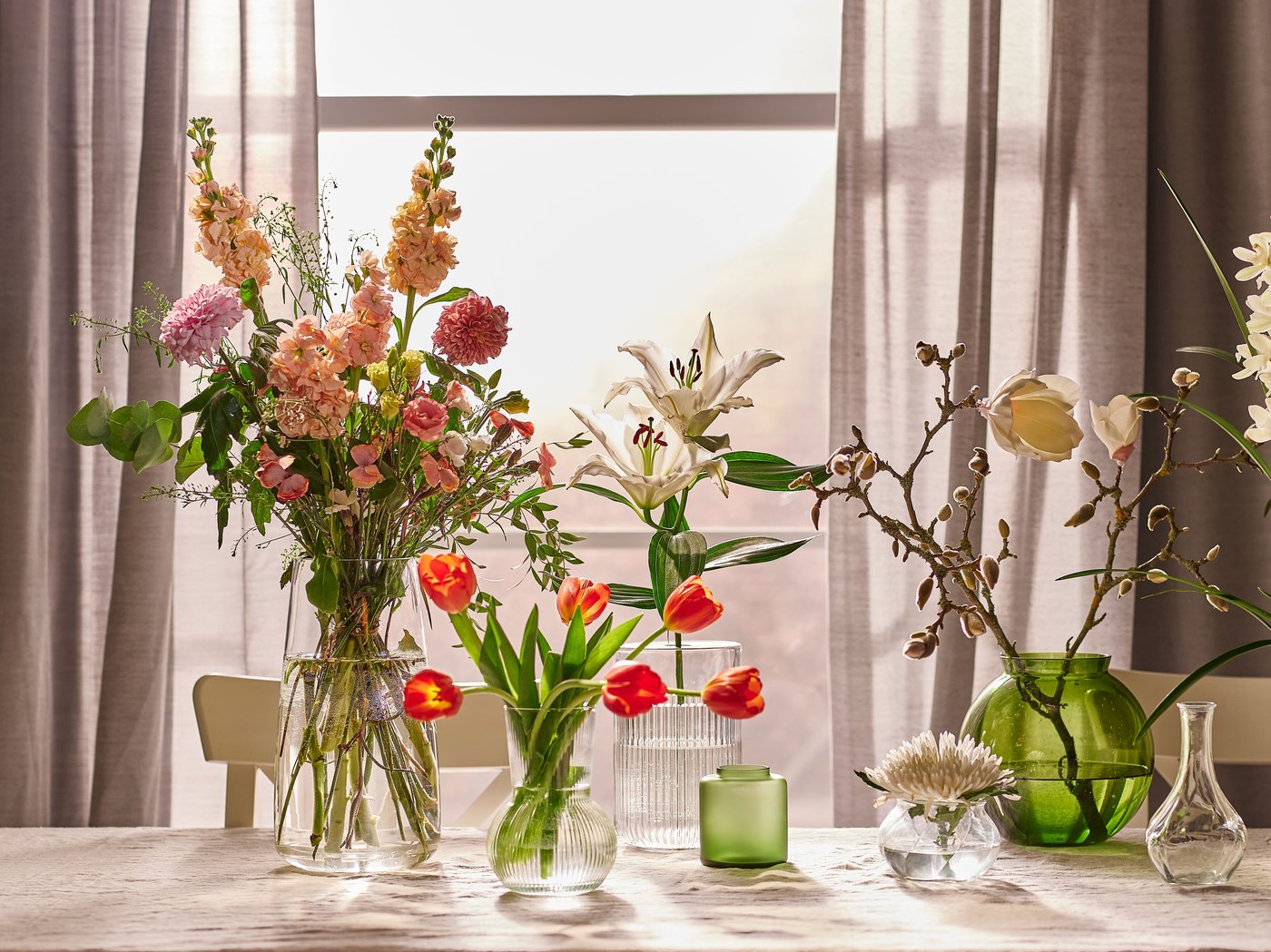 Es ist ein Tisch an einem Fenster mit Gardinenstores zu sehen. Auf dem Tisch sind verschiedene Blumenarrangements platziert.
