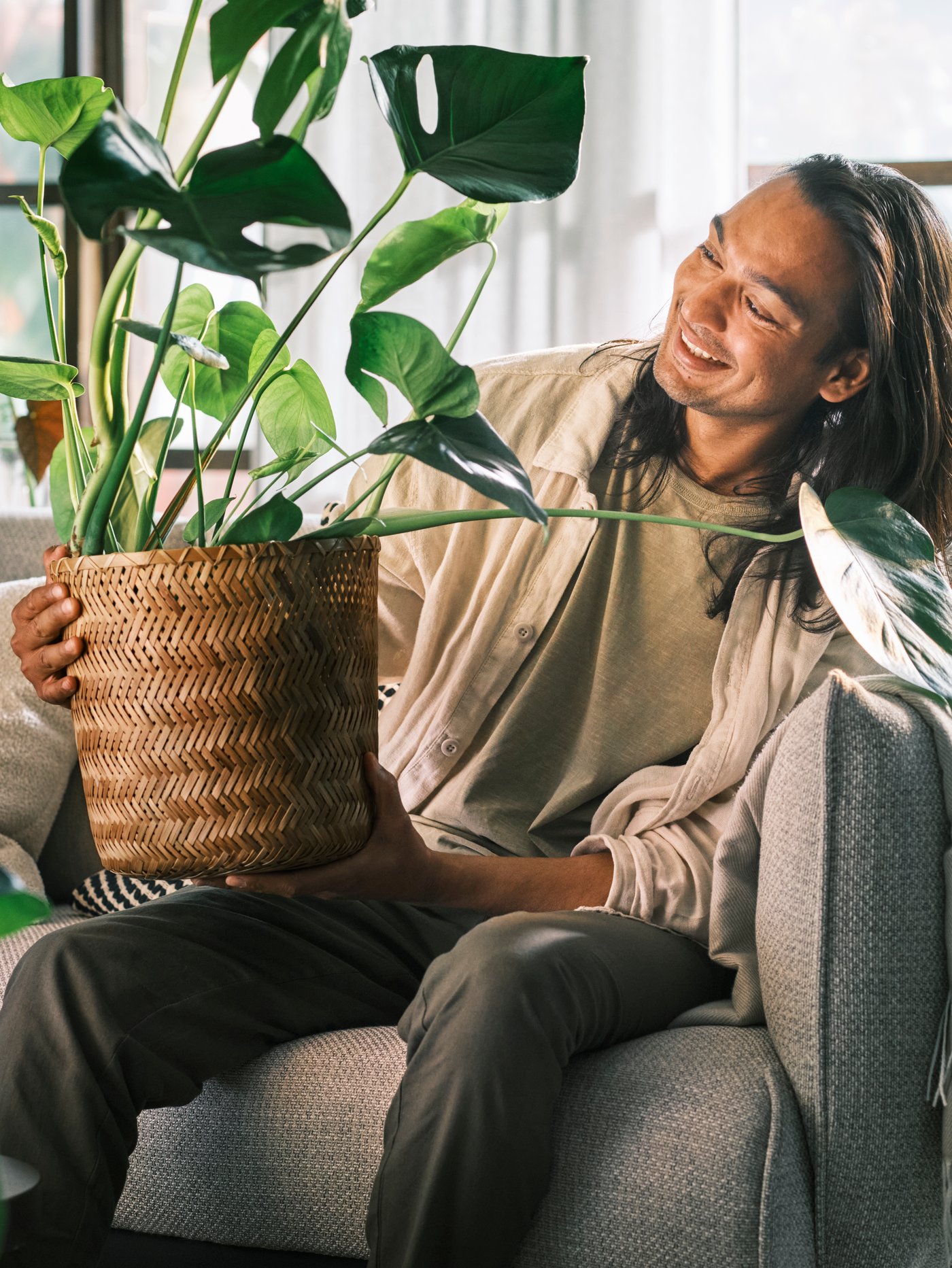 En person sidder i en lys sofa og kigger smilende på en plante i en håndlavet bambus KLYNON planteskjuler, som han holder på det ene knæ.