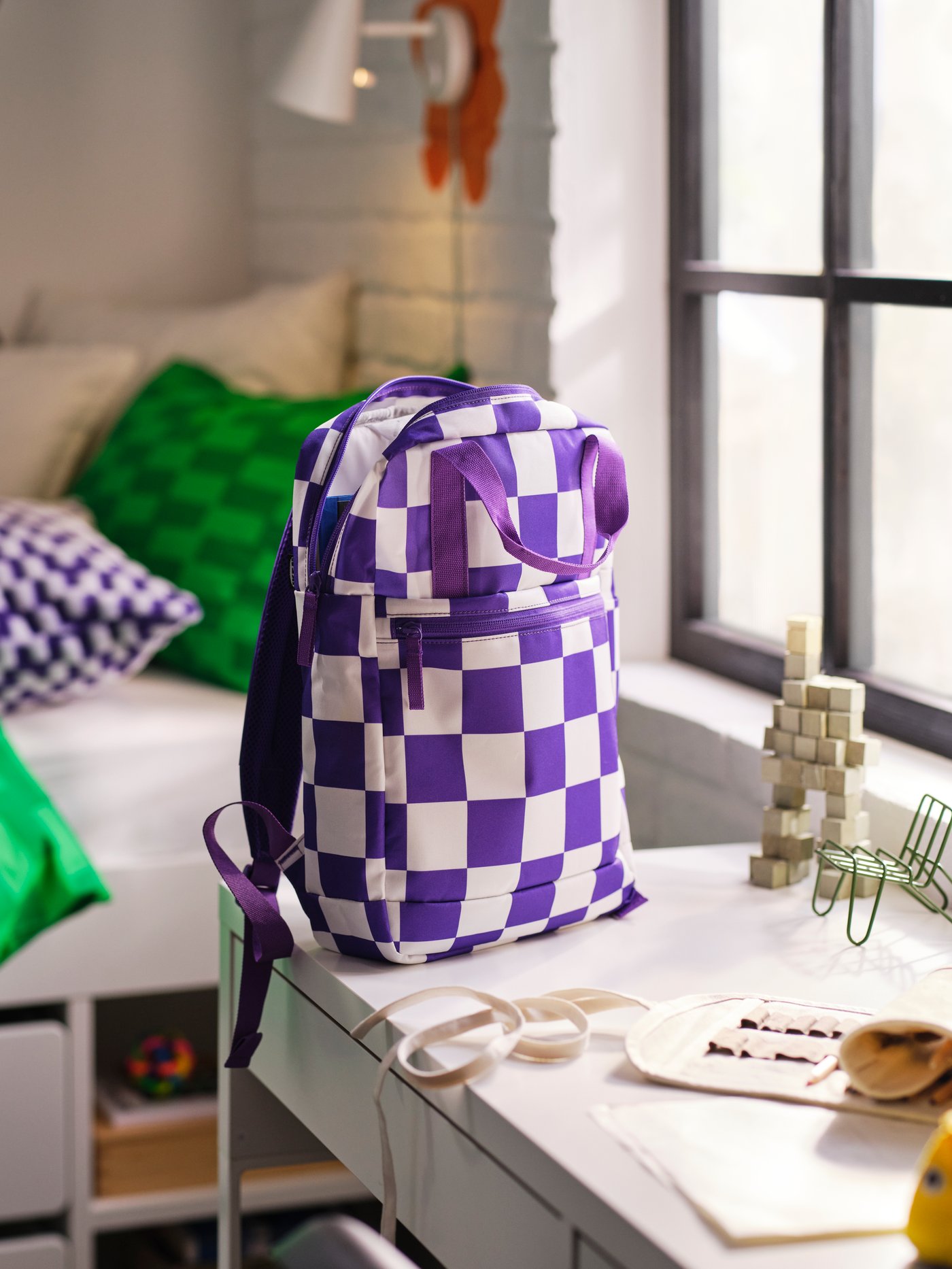 An IKEA STARTTID backpack in a white and purple check pattern on a white desk in a child room.


