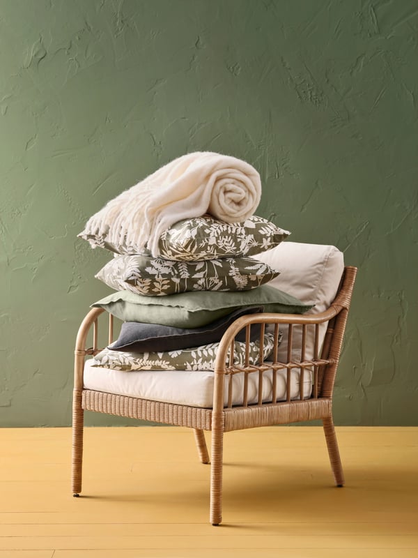 A HOLMSTA/FRÖKNABO rattan chair with a pile of ROTVECKLARE and DYTÅG cushions stacked in it. A white KLIPPOXEL throw on top.