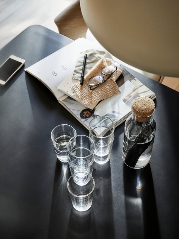 Glasses and a glass carafe with a cork stopper stand on a black table next to an open magazine and a smartphone.