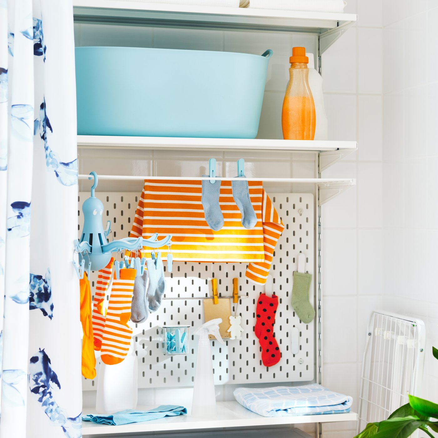 Repisas blancas BOAXEL instaladas a muro detrás de una cortina de baño blanca con patrones azules. Sobre las repisas se ven botellas con insumos de lavado y ropa de bebé roja y naranja colgada.