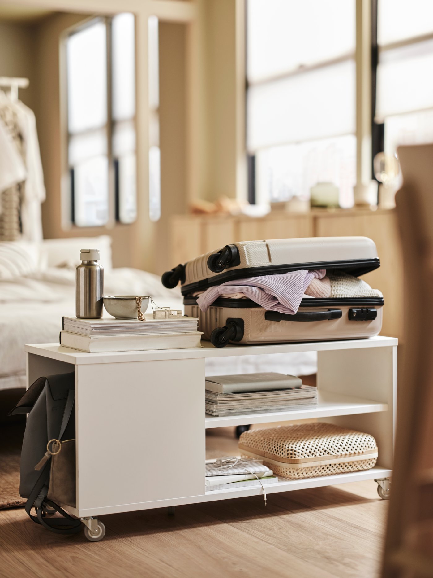 In a beige bedroom is a white VIHALS coffee table. On top of the table is a travel case. Books line the shelves underneath.