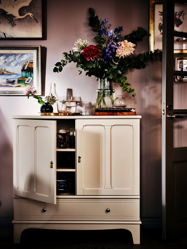 An IKEA LOMMARP off-white cabinet with gold handles and knobs in a room with dusty pink walls, flowers in a vase and various decoration on top.