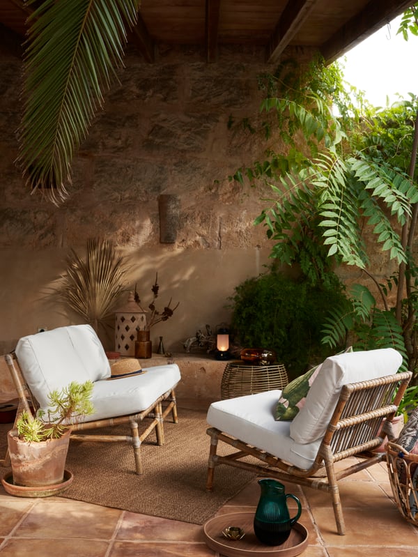 An outdoor setting showing two wicker TVARO chairs with white cushions, and green plants draped in the background.