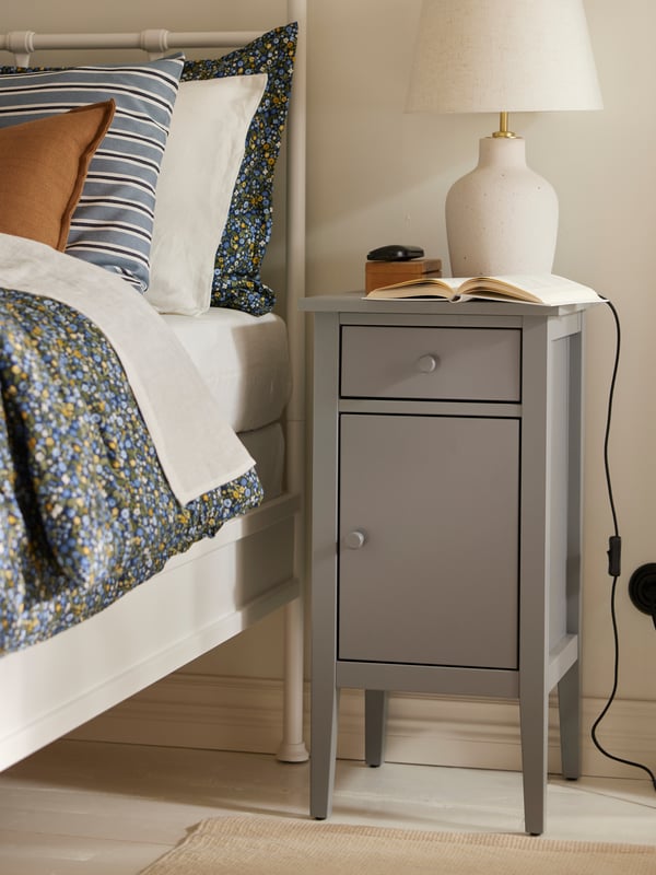 A grey GULLABERG bedside cabinet with an off white BILDVÄDER table lamp and an open book on it beside a white STJÄRNÖ bed.