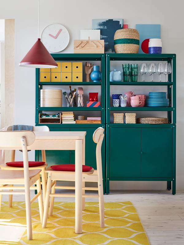 Table and chairs in birch, red chair pads, a yellow rug, a dark red pendant lamp and two green shelving units with cabinets.