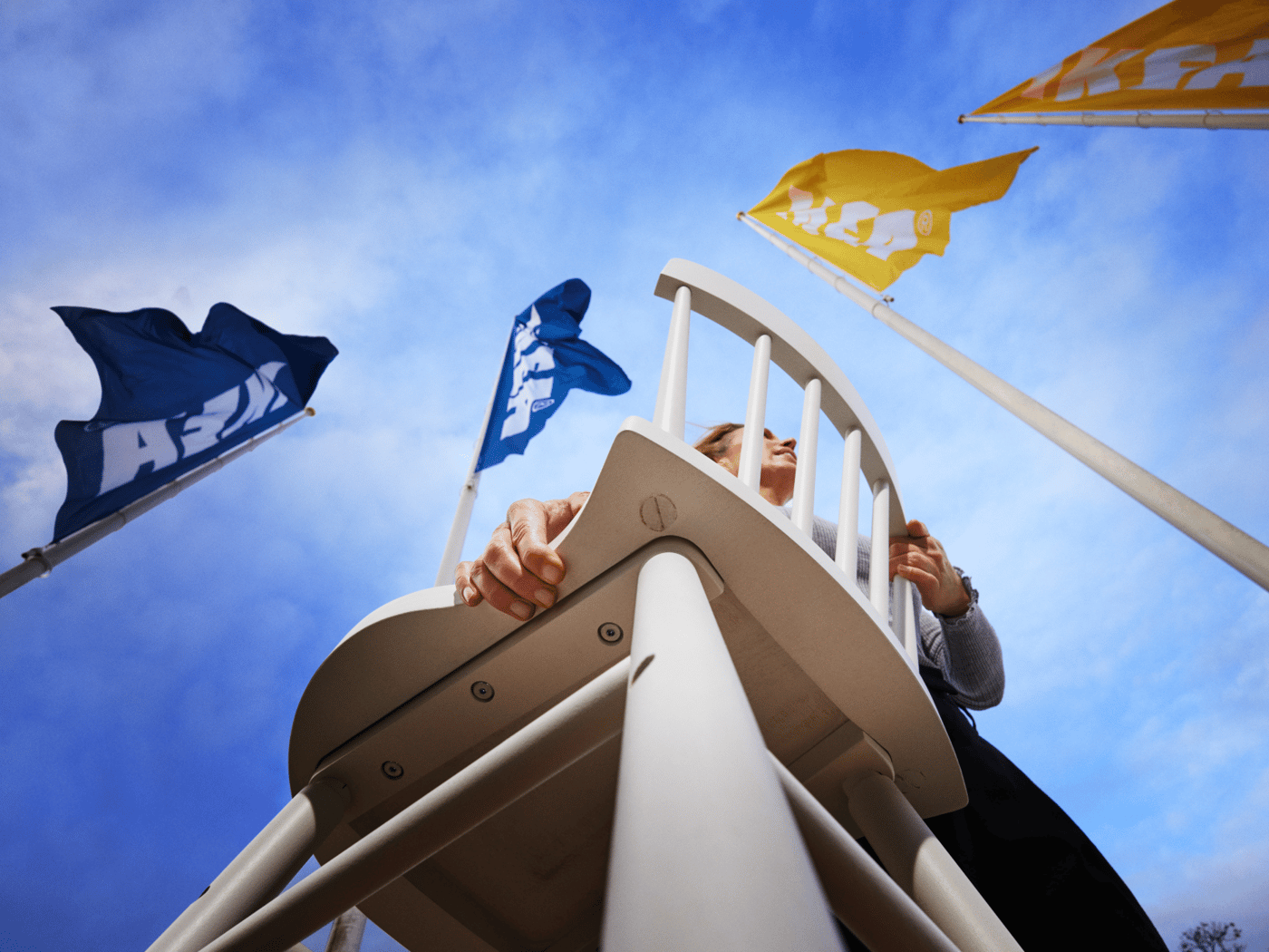 Person holding up and carrying a chair outdoors, photographed from below, with several flags waving against a blue sky.