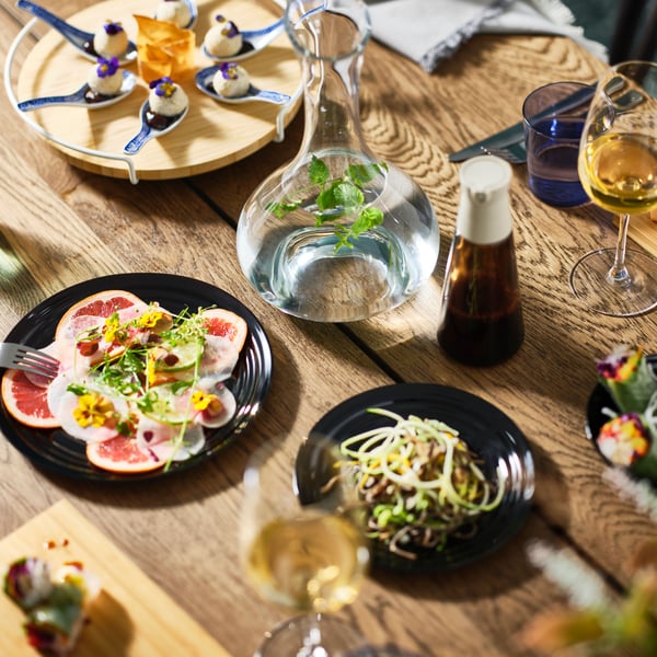 Et MÖRBYLÅNGA bord i mørk egetræsfiner er dækket med tallerkener i forskellige farvede og festlige fade samt en STORSINT karaffel.