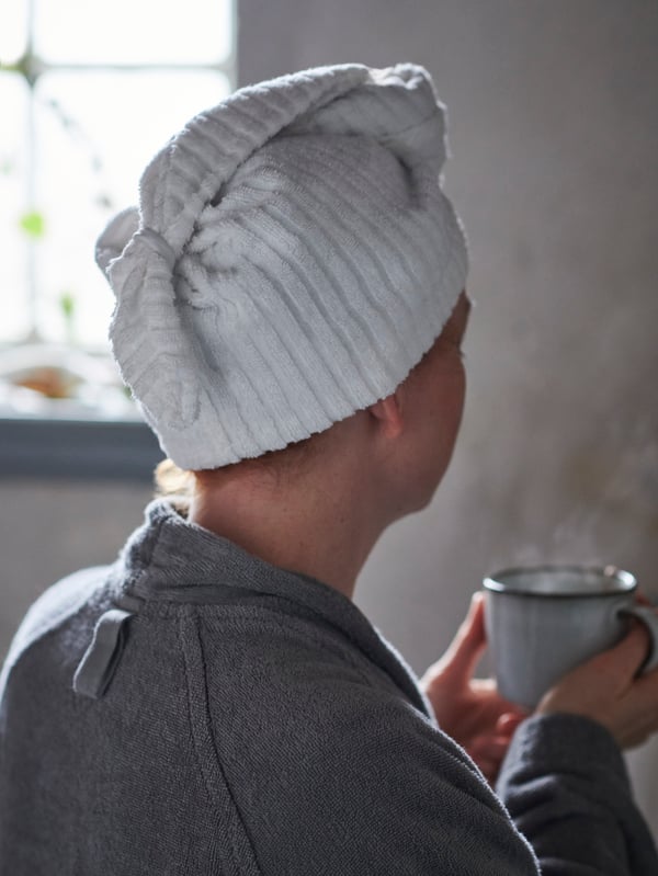 En person iklädd morgonrock och hårhandduk syns snett bakifrån hållandes en tekopp.