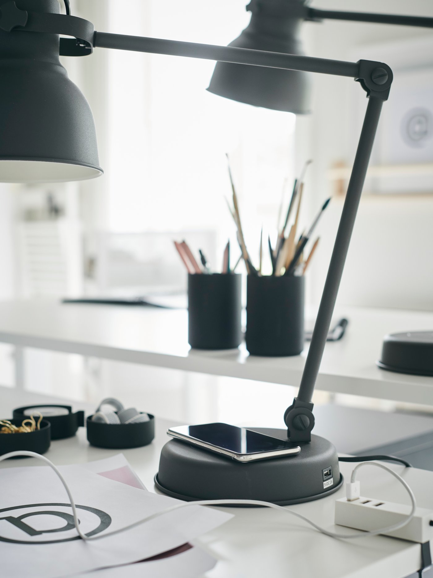 Eine Arbeitsleuchte auf einem Schreibtisch. Auf ihrem Standfuss liegt ein Mobiltelefon. Auf dem Tisch sind ein geschlossener Laptop, ein kleines Notizbrett und andere Bürogegenstände zu sehen.