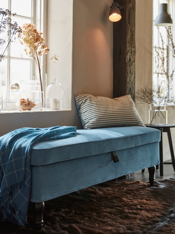 La esquina de una habitación con un sofá azul sobre una alfombra de piel. Detrás de el sofá una ventana con adornos florales en el bore