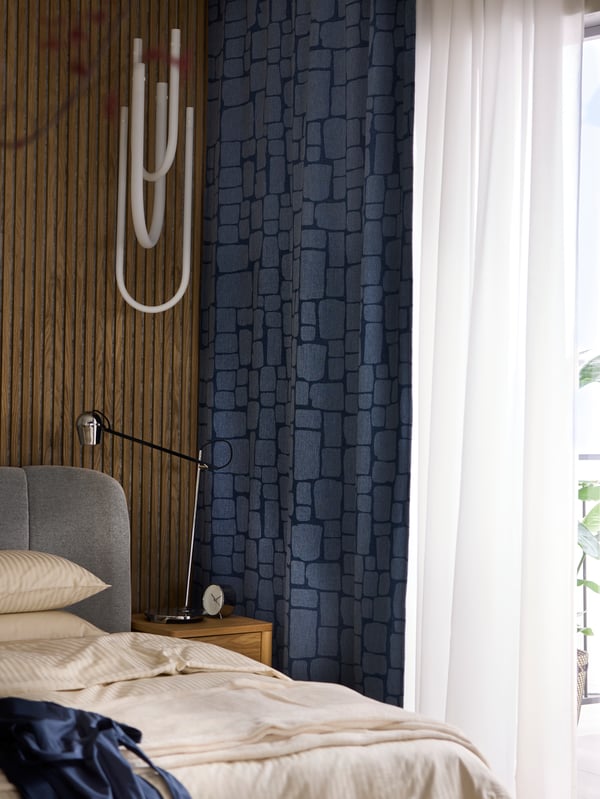 The corner of a bedroom by a window that has LÖNNSTÄVMAL block-out curtains and ROSENROBINIA sheer curtains hanging on it.