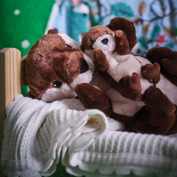 A SKOGSDUVA soft toy otter with baby is lying on a white GULSPARV blanket in a DUKTIG doll’s bed.