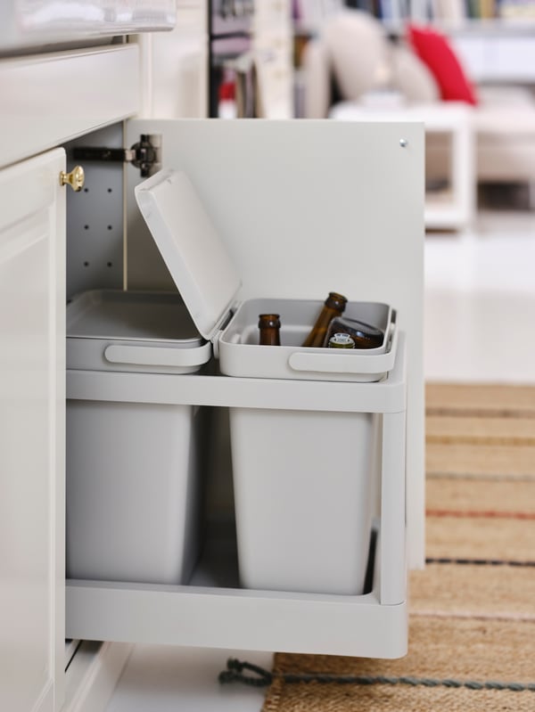 A pulled-out kitchen drawer revealing two HÅLLBAR bins in light grey. A bin for glass bottles has the lid open.