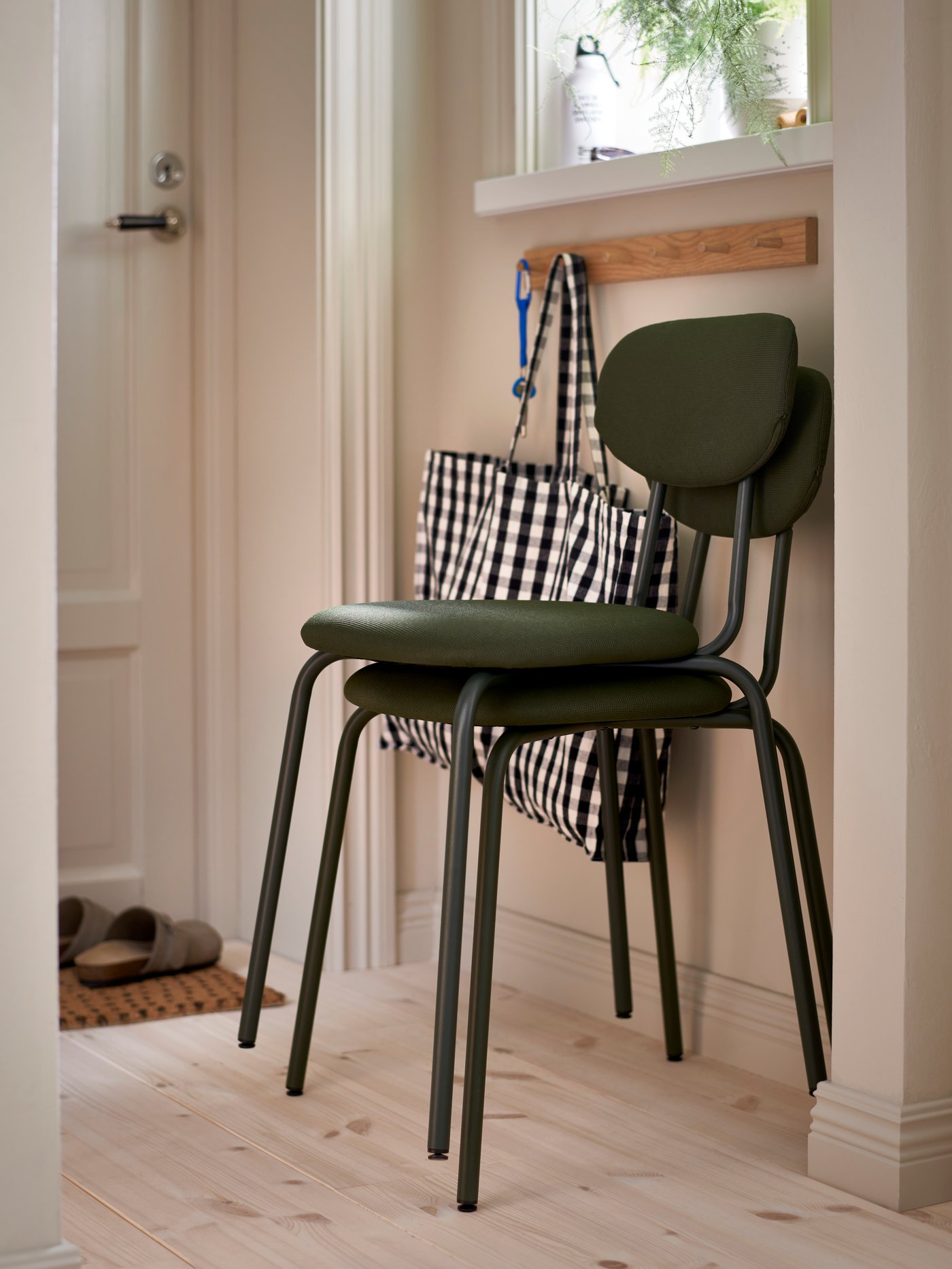 A deep green ÖSTANÖ chair is stacked on top of another identical chair under a window near a bag hanging from a knob.