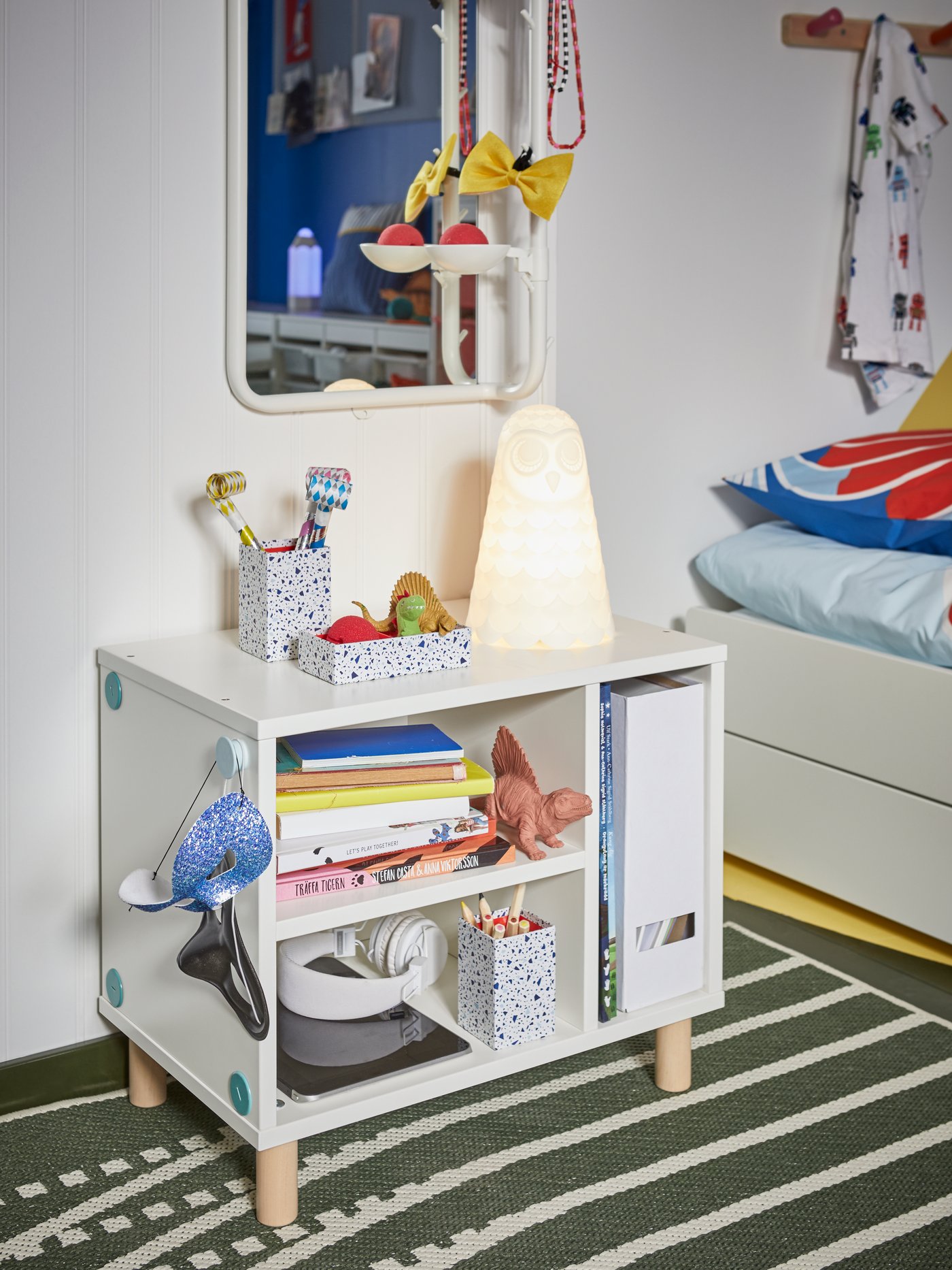 A white bedside table with open shelves showing books, pens, masks, headphones, dinosaur toys and an owl bedside lamp.