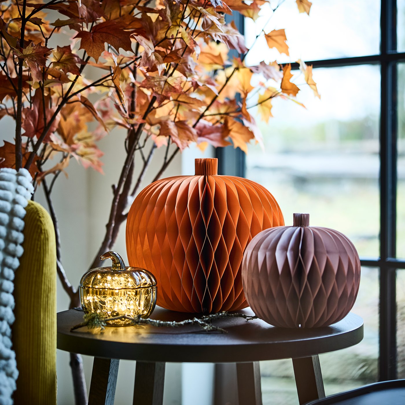 A set of two HÖSTAGILLE decoration paper pumpkins and a pumpkin-shaped bowl with lid are on a side table next to a window.