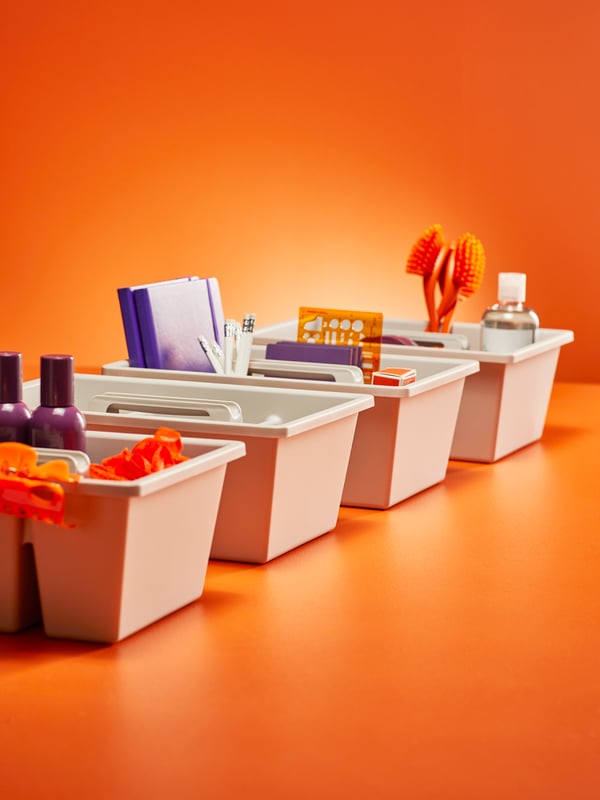Quatre plateaux à compartiments FÅNGGRÖDA contenant des produits ménagers, alignés avec soin dans un espace orange vif.