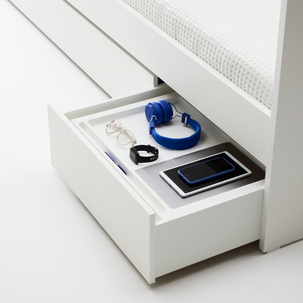 An open drawer underneath a white VIHALS daybed, showing storage for headphones, a phone, and accessories.