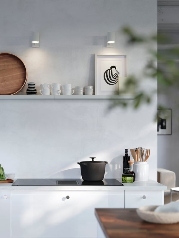 A white MOSSLUNDA picture ledge is placed on a wall above a kitchen stove and it has a picture frame, a tray and cups on it.