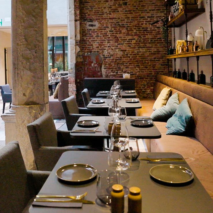 Rij gedekte tafels langs bakstenen muur met kussens en decoratie. Sfeervol restaurantinterieur met warme tinten en rustieke elementen.