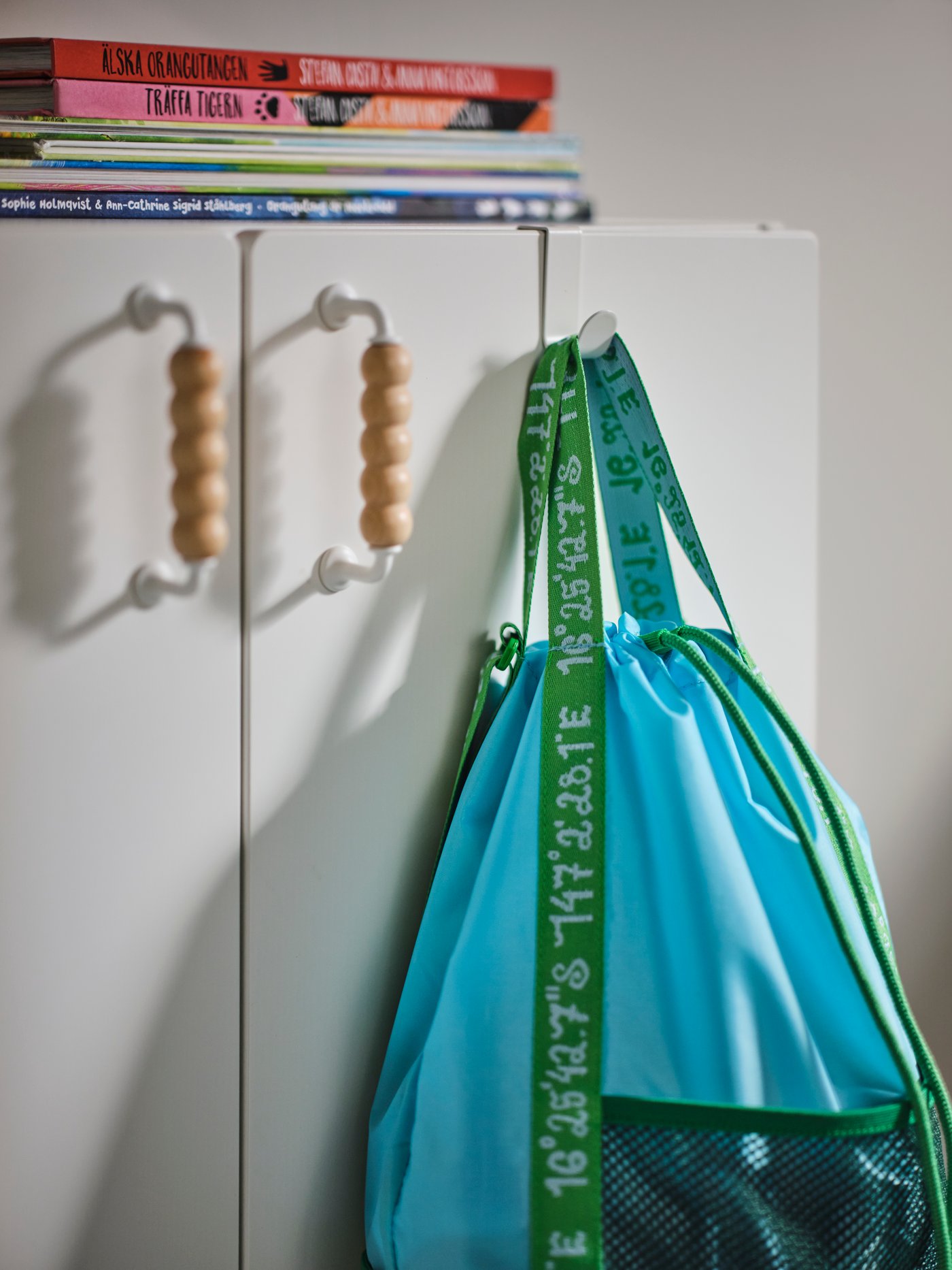 A white wall cabinet with books on top and a blue/green backpack on a white LÄTTHET hook and clip on one of the doors.