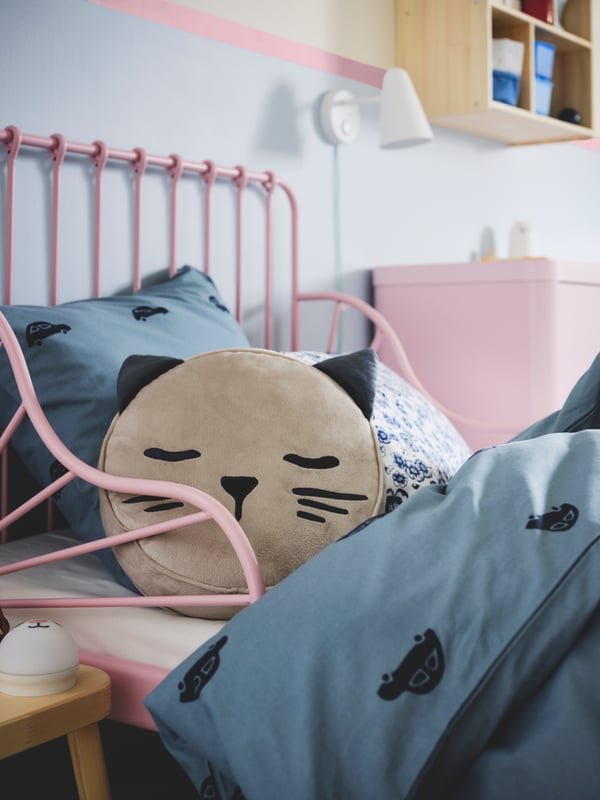 Ein Kinderzimmer mit einem beigefarbenen BARNDRÖM Kissen mit Tasche und einem Katzengesicht auf einem ausziehbaren Bett in Hellrosa.