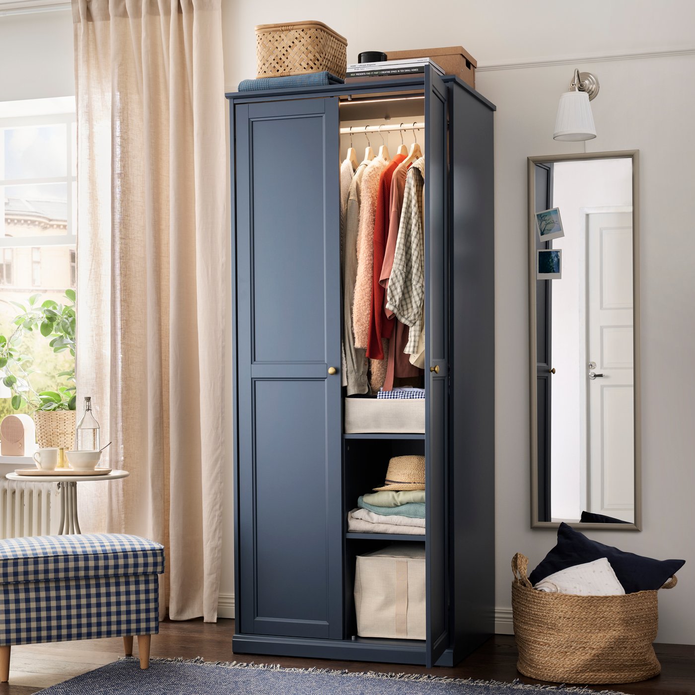 A bedroom with a blue FÅGELFJÄLLET wardrobe displaying clothes inside and a mirror and window in the background.