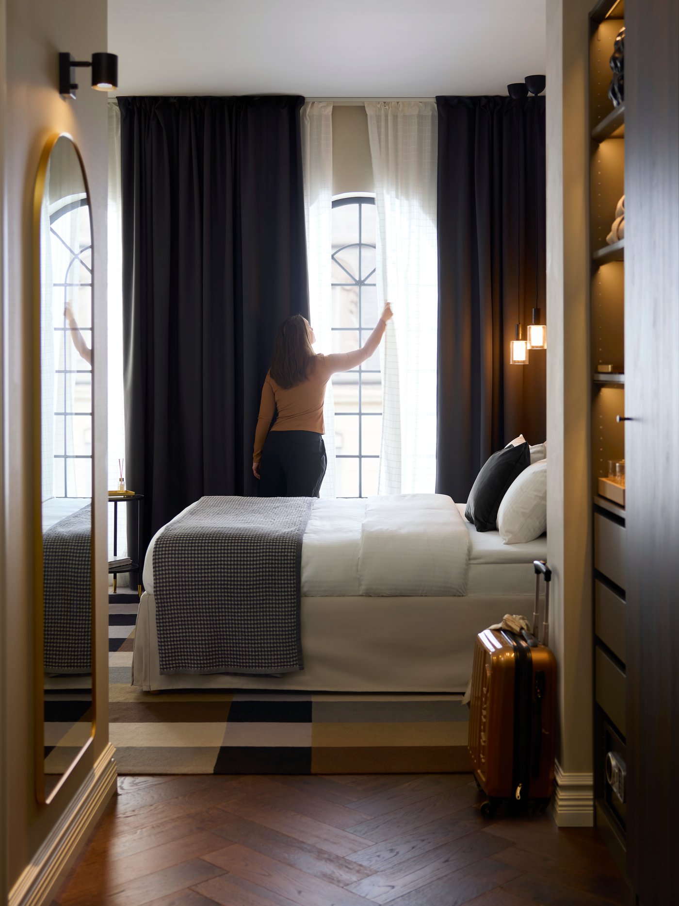 A person is by dark grey KORGMOTT block-out curtains that partially cover sheer curtains on a large, sunny hotel room window.