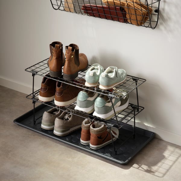 Two GREJIG shoe racks in black metal stacked together holding trainers and boots on a tray in a hallway.
