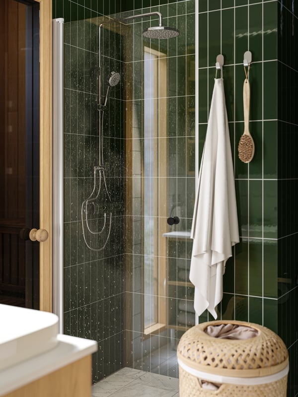 A shower is placed in a green-tiled bathroom and there is a white FREDIKSJÖN towel hanging from hooks on the wall next to it.