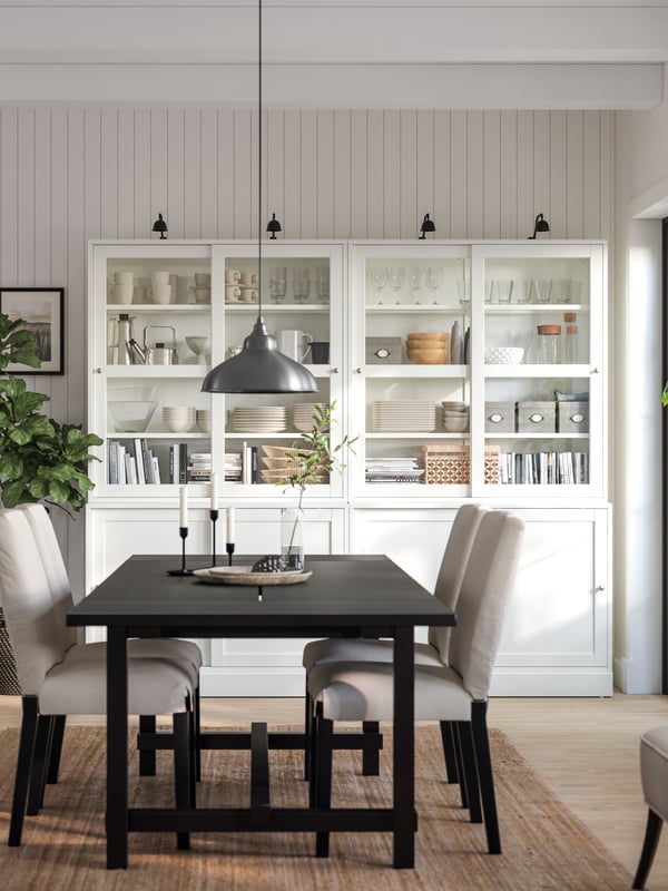 A classic dining room with a black dining table surrounded by four upholstered chairs. Against the wall stands a white HAVSTA sideboard displaying tableware and glassware.
