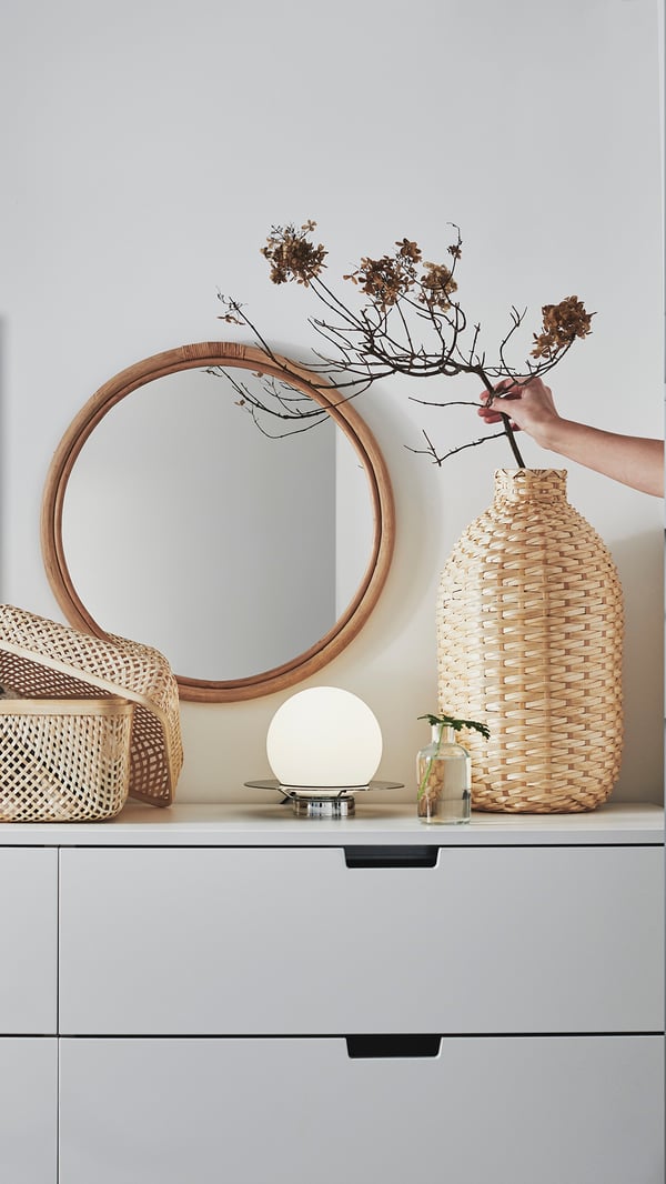 Neutral small homewares sit on a modern chest of drawers, a small round mirror sits behind thme with a wicker vase
