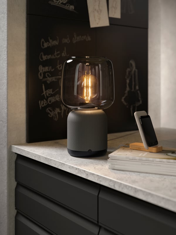 Un chargeur sans fil NORDMÄRKE sur un comptoir près d'un haut-parleur lampe de table SYMFONISK.