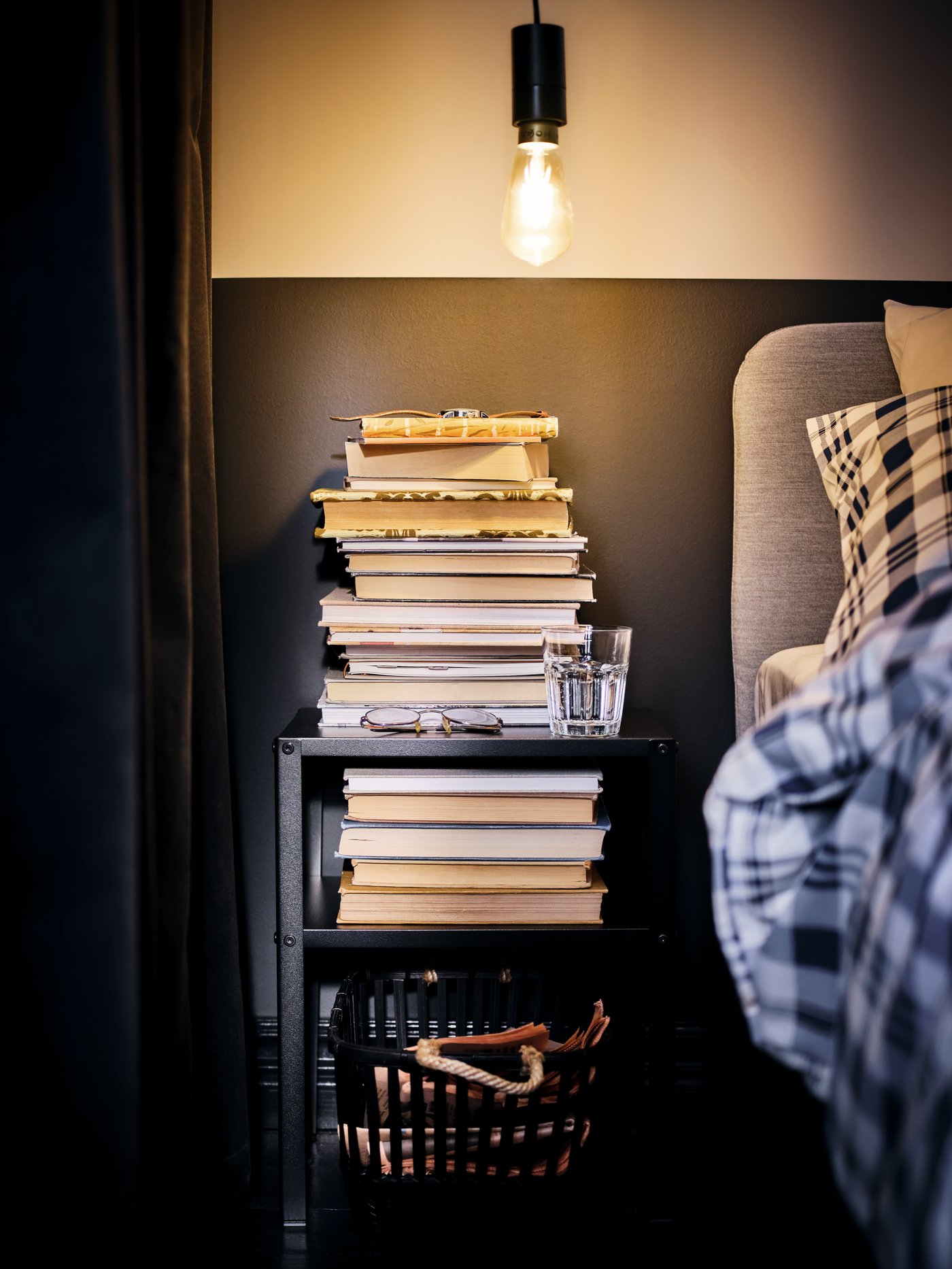 Une ampoule nue suspendue illumine un lit et une table de chevet KNARREVIK noire sur laquelle beaucoup de livres sont empilés.