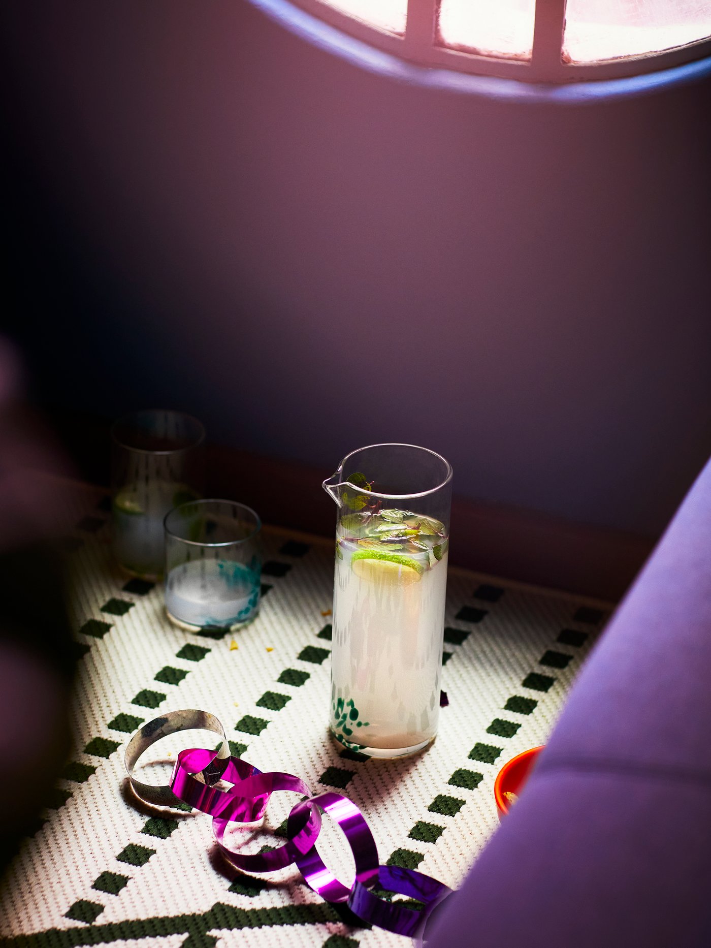 An ÖMSESIDIG carafe in clear and green glass filled with lemonade, next to two ÖMSESIDIG glasses in different sizes.