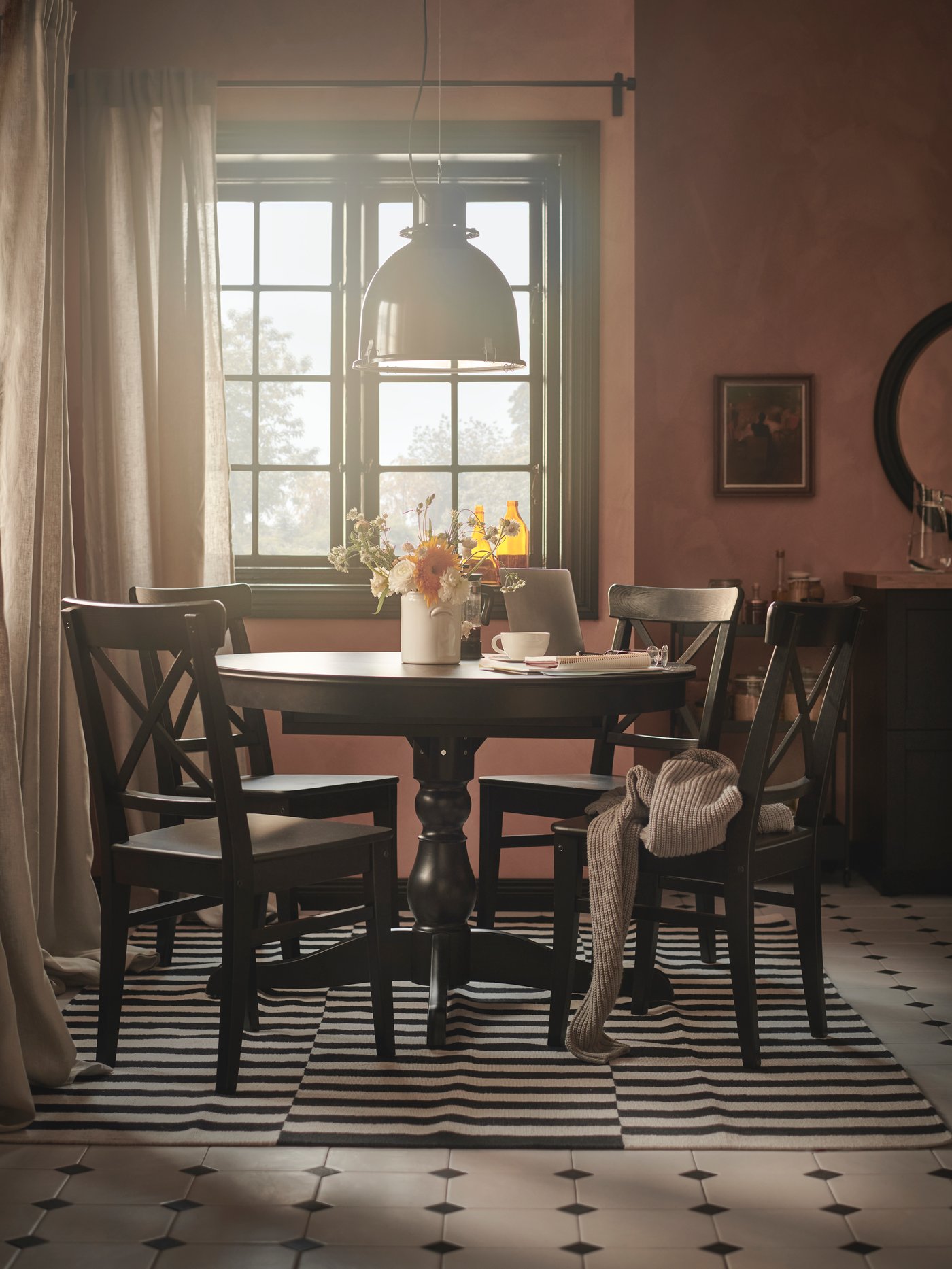 Runt träbord med fyra stolar står dukat med te och blommor i ett varmt upplyst rum med fönster, matta och dekorativt sideboard.
