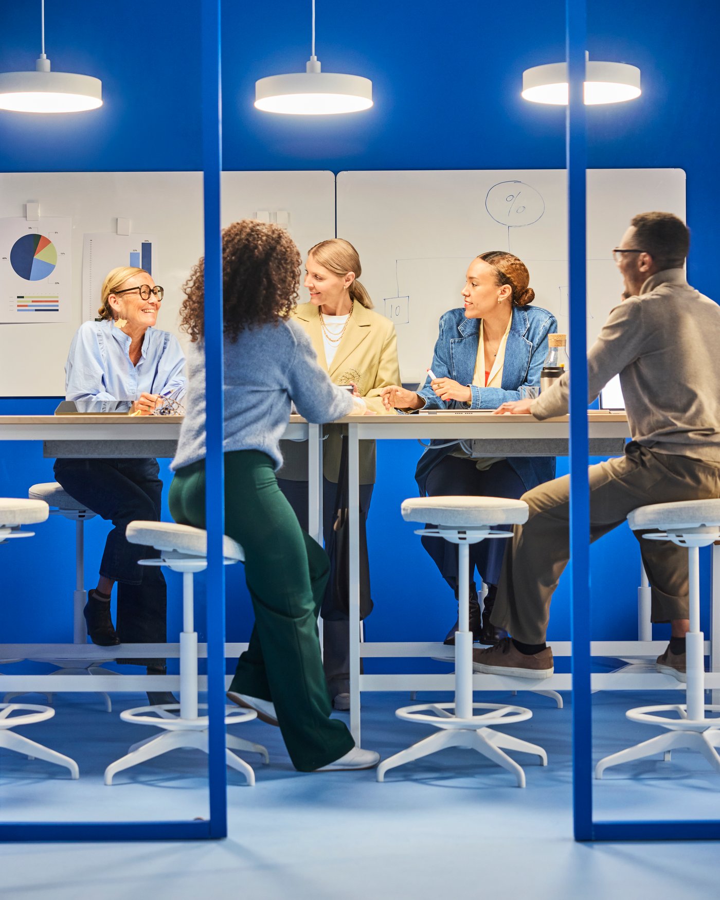 An office setting with a group of meeting gathered at a hightop desk sitting on stools.