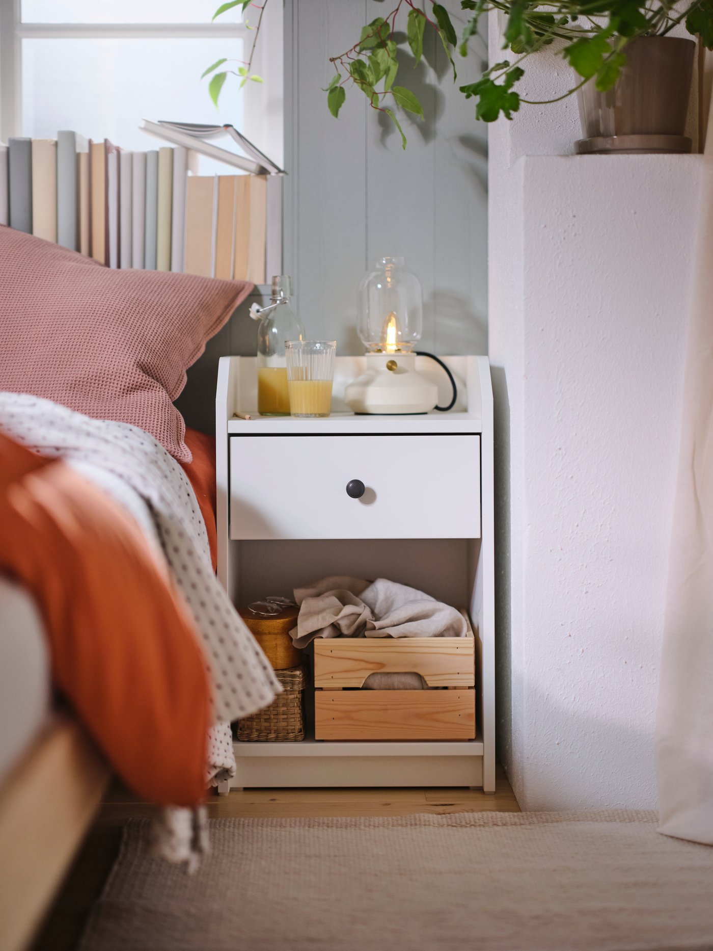A white HAUGA bedside table with a TÄRNBY table lamp on top. A cushion in a pale pink KLOTSTARR cover is on the bed by it.