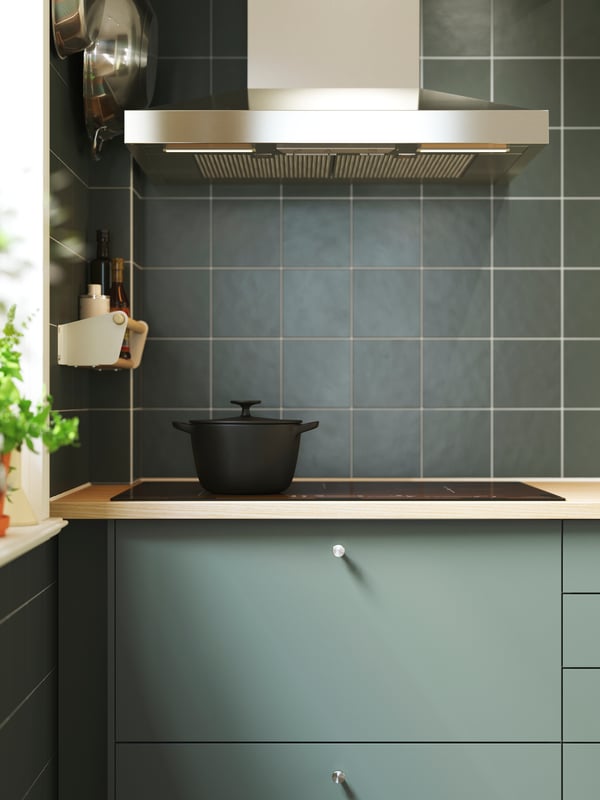 Cuisine carrelée de vert équipée d’armoires avec façades NICKEBO gris vert, d’une hotte et d’une table de cuisson noire sur laquelle est posée une cocotte en fonte.