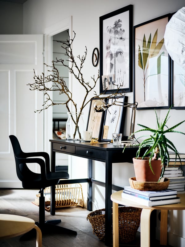 A HEMNES desk with a vase of branches on top, against a white wall covered with pictures. A swivel chair is in front.
