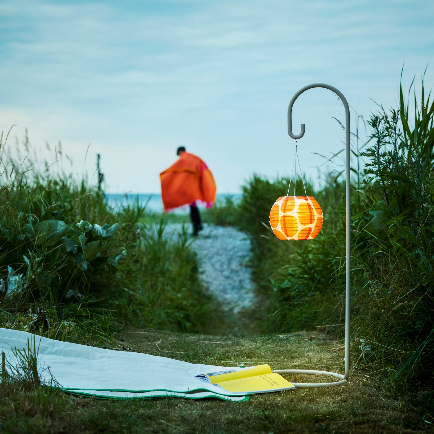 Un escenario nocturno en el jardín muestra un soporte para lámparas SOLVINDEN beige, con una pantalla naranja iluminada y una persona caminando.