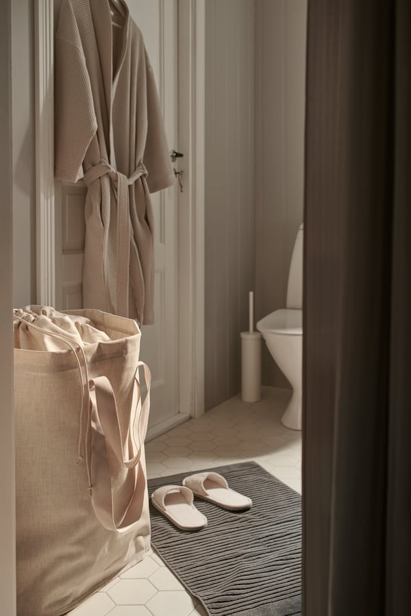 Vue latérale d’une salle de bains dans laquelle se trouvent un sac à lessive PURRPINGLA beige et un peignoir beige accroché à un crochet.