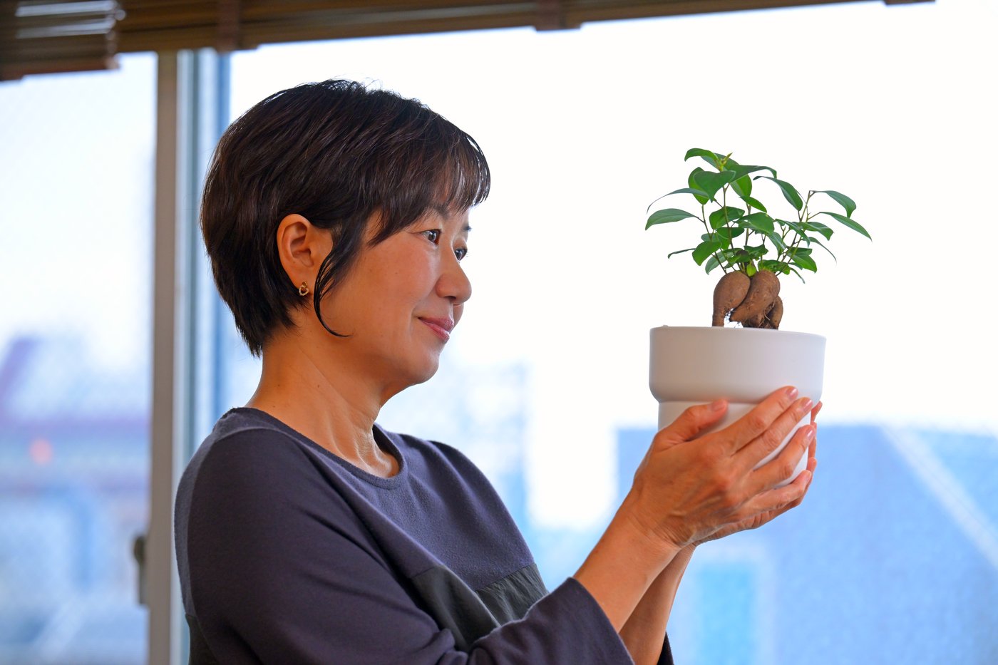 Žena drži belu saksiju u kojoj je malo bonsai drvo.