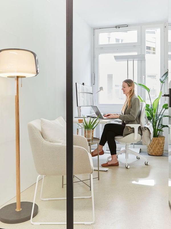 In einem Büro mit Glasfront sitzt eine Frau vor dem Fenster an einem Schreibtisch. Neben ihr steht eine Topfpflanze und davor ein Sessel für Besucher mit Beistelltisch und dahinter eine Stehlampe aus Glas mit einem Fuss aus Holz und Marmor. 