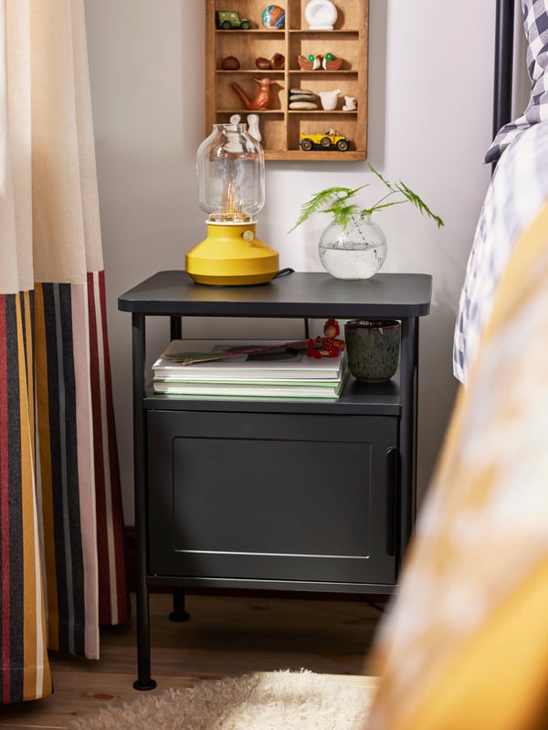Close-up of GRÅFJÄLLET bedside table in anthracite with yellow TÄRNABY table lamp and small glass vase on top.